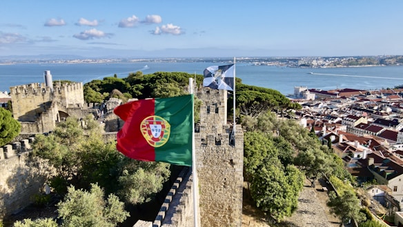 A historic stone castle overlooking a scenic bay with lush green trees. The Portuguese flag flutters in the wind atop the castle walls. In the background, there is a panoramic view of a coastal city with red roofed buildings and a serene blue ocean.