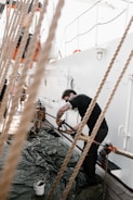 A person with tattoos on their arm is engaged in a task on a boat deck. They are leaning over ropes and metal parts, possibly working or cleaning. The deck has a tarp spread out, with a paint bucket nearby. Bright lighting suggests it is daytime.