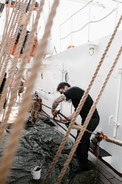 A person with tattoos on their arm is engaged in a task on a boat deck. They are leaning over ropes and metal parts, possibly working or cleaning. The deck has a tarp spread out, with a paint bucket nearby. Bright lighting suggests it is daytime.