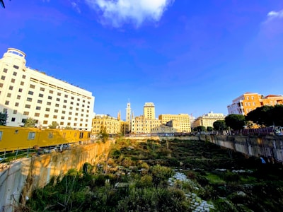 A view of a construction site surrounded by various buildings. The foreground shows a large pit with overgrown vegetation and remnants of old structures, indicating an area under development or excavation. The background features a mix of modern and classical buildings under a clear blue sky.