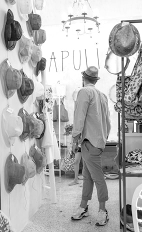 A person wearing a hat and casual clothing stands in a store that has numerous hats displayed on the wall. The interior is stylish with a chandelier and the word 'APULIA' is visible in large letters. Hats and other accessories are neatly arranged on racks and shelves.