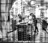 A bustling kitchen with a chef preparing dishes, captured in black and white. The scene is viewed through a gridded window, which adds a blurred, artistic effect. Stainless steel equipment, countertops, and shelves fill the background, along with stacks of plates.
