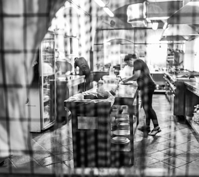 A bustling kitchen with a chef preparing dishes, captured in black and white. The scene is viewed through a gridded window, which adds a blurred, artistic effect. Stainless steel equipment, countertops, and shelves fill the background, along with stacks of plates.