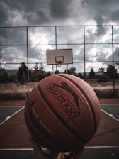 Close-up of a hand holding a Lift Athletics Inc basketball on an outdoor court at sunset.