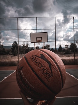 A basketball is being held close to the camera, with an outdoor court and hoop visible in the background. The sky is overcast with dark clouds, giving a dramatic atmosphere.