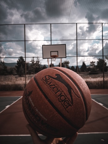 Close-up of a hand holding a Lift Athletics Inc basketball on an outdoor court at sunset.