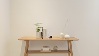 A minimalist wooden side table with clean lines, set against a neutral-toned wall with a small green plant beside it.