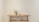 A wooden table is positioned against a plain, light-colored wall. On top of the table, there is a small decorative plant in a pot, several books standing upright, and two minimalistic vases. An additional small, black object is placed beside the books. The lower shelf holds a few stacked items, including a geometric sculpture.