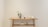 A wooden table is positioned against a plain, light-colored wall. On top of the table, there is a small decorative plant in a pot, several books standing upright, and two minimalistic vases. An additional small, black object is placed beside the books. The lower shelf holds a few stacked items, including a geometric sculpture.