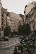 A European city street lined with vintage-style buildings featuring ornate architecture. A curved road cuts through the center, with parked cars along its sides and a cluster of bicycles in the foreground. The area is serene with minimal foot traffic, and trees add greenery to the urban setting.
