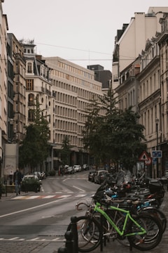 A European city street lined with vintage-style buildings featuring ornate architecture. A curved road cuts through the center, with parked cars along its sides and a cluster of bicycles in the foreground. The area is serene with minimal foot traffic, and trees add greenery to the urban setting.