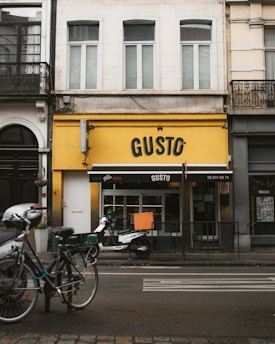 A street view of a building with a bright yellow facade and the name 'GUSTO' displayed prominently. Two bicycles and a scooter are parked in front, and a black awning with white text covers the entrance. The building's upper section has white and cream walls with shuttered windows.