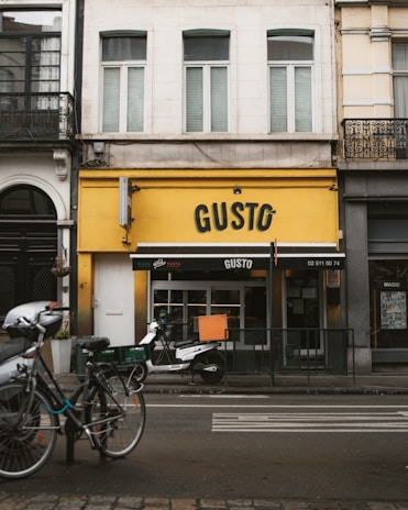 A street view of a building with a bright yellow facade and the name 'GUSTO' displayed prominently. Two bicycles and a scooter are parked in front, and a black awning with white text covers the entrance. The building's upper section has white and cream walls with shuttered windows.