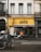 A street view of a building with a bright yellow facade and the name 'GUSTO' displayed prominently. Two bicycles and a scooter are parked in front, and a black awning with white text covers the entrance. The building's upper section has white and cream walls with shuttered windows.
