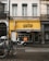 A street view of a building with a bright yellow facade and the name 'GUSTO' displayed prominently. Two bicycles and a scooter are parked in front, and a black awning with white text covers the entrance. The building's upper section has white and cream walls with shuttered windows.