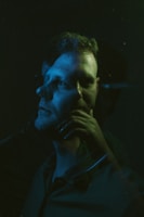 A dramatic double exposure portrait featuring a person with their profile overlapping a front-facing angle. The image is illuminated with moody blue and green lighting, creating a surreal and introspective atmosphere.