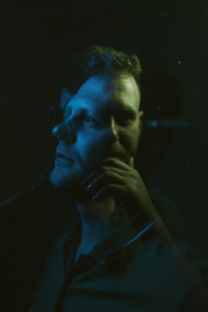 A dramatic double exposure portrait featuring a person with their profile overlapping a front-facing angle. The image is illuminated with moody blue and green lighting, creating a surreal and introspective atmosphere.