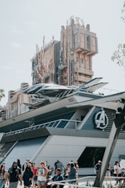 A futuristic building with elaborate architectural details, resembling a theme park attraction. The structure features a prominent logo and metallic elements, with a spacecraft-like design attached to the side. People are gathered around the area, some walking and others taking photos.