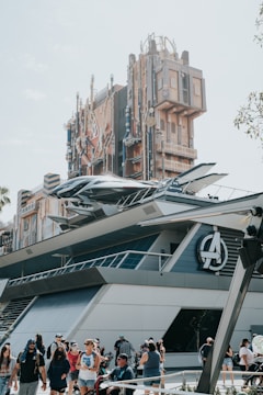 A futuristic building with elaborate architectural details, resembling a theme park attraction. The structure features a prominent logo and metallic elements, with a spacecraft-like design attached to the side. People are gathered around the area, some walking and others taking photos.
