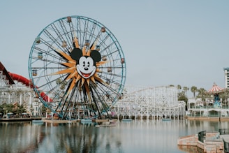 A large Ferris wheel with a central Mickey Mouse face stands near a body of water. The amusement park setting includes a roller coaster, various attractions, and landscaping with palm trees. The sky is clear, and the overall atmosphere is inviting and fun.