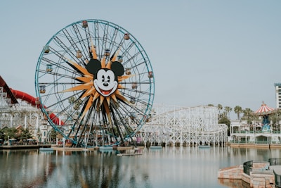 A large Ferris wheel with a central Mickey Mouse face stands near a body of water. The amusement park setting includes a roller coaster, various attractions, and landscaping with palm trees. The sky is clear, and the overall atmosphere is inviting and fun.