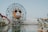 A large Ferris wheel with a central Mickey Mouse face stands near a body of water. The amusement park setting includes a roller coaster, various attractions, and landscaping with palm trees. The sky is clear, and the overall atmosphere is inviting and fun.