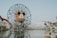 A large Ferris wheel with a central Mickey Mouse face stands near a body of water. The amusement park setting includes a roller coaster, various attractions, and landscaping with palm trees. The sky is clear, and the overall atmosphere is inviting and fun.