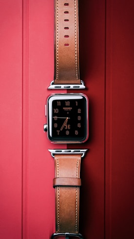 A smartwatch with a square face and digital display is elegantly designed with a leather strap. It is positioned vertically against a red background, highlighting the luxurious appeal of the watch.