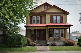 A two-story house with beige siding and maroon trim, featuring a front porch supported by wooden columns. There is a lush green tree on the left side and small bushes flanking the pathway leading to the entrance.