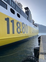 A large ferry with a prominent yellow and black color scheme is docked at a pier. The number 11880 is clearly visible on the side of the vessel, along with several windows and lifeboats on the upper deck. The water in the harbor is calm, with a mountainous landscape visible in the distance under a clear sky.