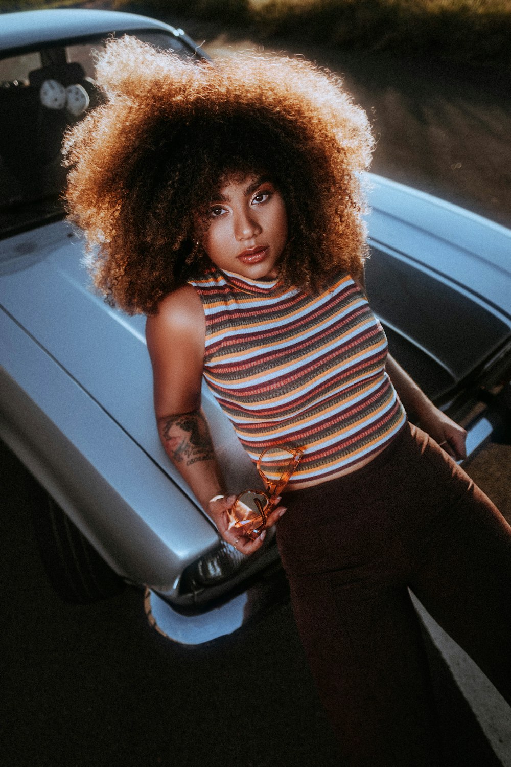 girl in white and black stripe tank top and black pants standing beside gray car