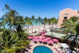 A tropical beach scene with palm trees surrounding a large building. The image features a bustling beach area with numerous sun umbrellas and lounge chairs. The ocean is a vivid blue, and there is a swimming pool surrounded by pink sun umbrellas and greenery in the foreground.
