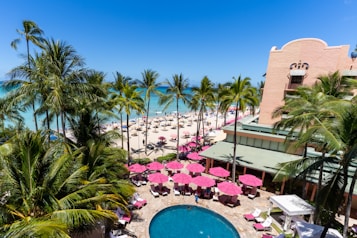 A tropical beach scene with palm trees surrounding a large building. The image features a bustling beach area with numerous sun umbrellas and lounge chairs. The ocean is a vivid blue, and there is a swimming pool surrounded by pink sun umbrellas and greenery in the foreground.