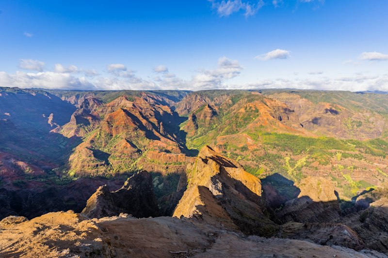Wide canyon of red and orange ridges streaked with green vegetation under a bright blue sky