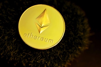 A golden coin featuring the Ethereum logo and text rests against a dark, textured surface. The lighting creates a dramatic, luxurious feel with reflective highlights on the coin.