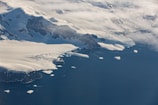 An aerial view of a large, snow-covered mountainous landscape with glaciers extending into a deep blue sea. Several icebergs are floating near the coastline, and patches of clouds partially obscure parts of the terrain.