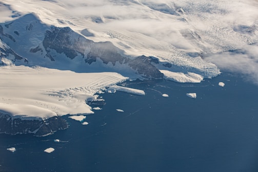 An aerial view of a large, snow-covered mountainous landscape with glaciers extending into a deep blue sea. Several icebergs are floating near the coastline, and patches of clouds partially obscure parts of the terrain.