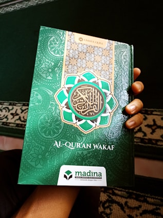 A hand holds a book with a decorative green cover displaying Arabic script and patterns. The book cover includes the title 'Al-Qur'an Wakaf' along with the branding of 'Madina'. There is a combination of ornate designs in white, gold, and green creating an intricate frame around the central text.