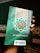 A hand holds a book with a decorative green cover displaying Arabic script and patterns. The book cover includes the title 'Al-Qur'an Wakaf' along with the branding of 'Madina'. There is a combination of ornate designs in white, gold, and green creating an intricate frame around the central text.