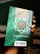 A hand holds a book with a decorative green cover displaying Arabic script and patterns. The book cover includes the title 'Al-Qur'an Wakaf' along with the branding of 'Madina'. There is a combination of ornate designs in white, gold, and green creating an intricate frame around the central text.