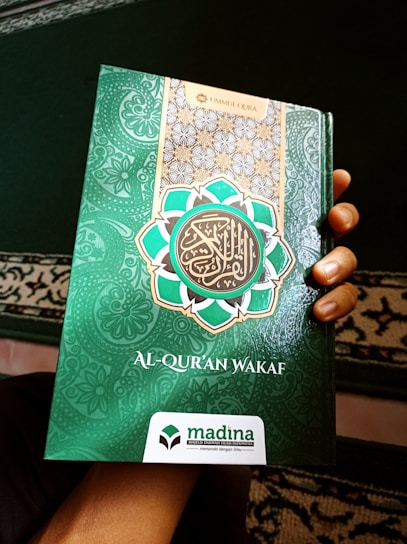 A hand holds a book with a decorative green cover displaying Arabic script and patterns. The book cover includes the title 'Al-Qur'an Wakaf' along with the branding of 'Madina'. There is a combination of ornate designs in white, gold, and green creating an intricate frame around the central text.