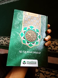 A hand holds a book with a decorative green cover displaying Arabic script and patterns. The book cover includes the title 'Al-Qur'an Wakaf' along with the branding of 'Madina'. There is a combination of ornate designs in white, gold, and green creating an intricate frame around the central text.