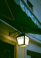 A hanging lantern displaying the name 'Ribot' twice on its illuminated surface, along with the word 'restaurant'. The lantern is attached to a building with a green awning that also bears the name 'Ribot'. The setting appears to be the exterior of a building with a partial view of a window and a door.