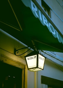 A hanging lantern displaying the name 'Ribot' twice on its illuminated surface, along with the word 'restaurant'. The lantern is attached to a building with a green awning that also bears the name 'Ribot'. The setting appears to be the exterior of a building with a partial view of a window and a door.