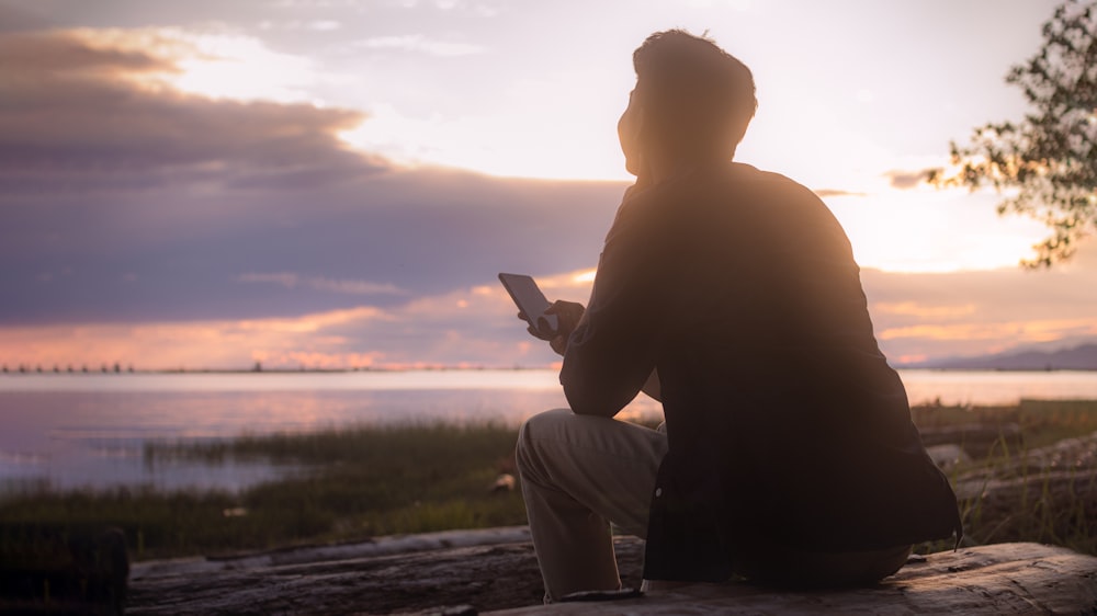 man in brown pants sitting on ground while holding smartphone during sunset