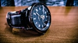 A close-up of a black wristwatch with Roman numerals on a brown wooden surface. The watch has a leather strap and intricate detailing on the dial with branding visible.