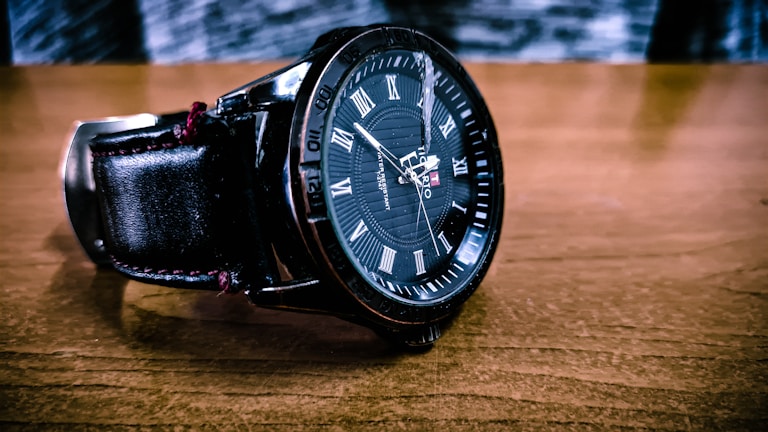 A close-up of a black wristwatch with Roman numerals on a brown wooden surface. The watch has a leather strap and intricate detailing on the dial with branding visible.