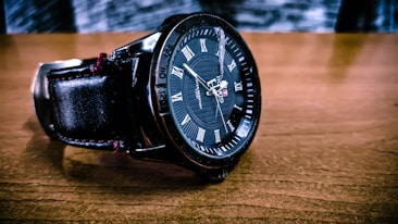 A close-up of a black wristwatch with Roman numerals on a brown wooden surface. The watch has a leather strap and intricate detailing on the dial with branding visible.