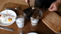 A person is pouring coffee from a cezve into a glass through a metal strainer. The table is set with a plate of fried eggs and sliced tomatoes, a cutting board, and various utensils.
