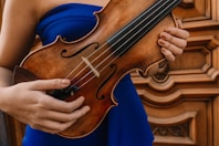 A person wearing a blue garment holds a brown wooden violin, with intricate woodwork in the background. The hands are positioned to hold and play the instrument, showcasing both the body and strings of the violin.